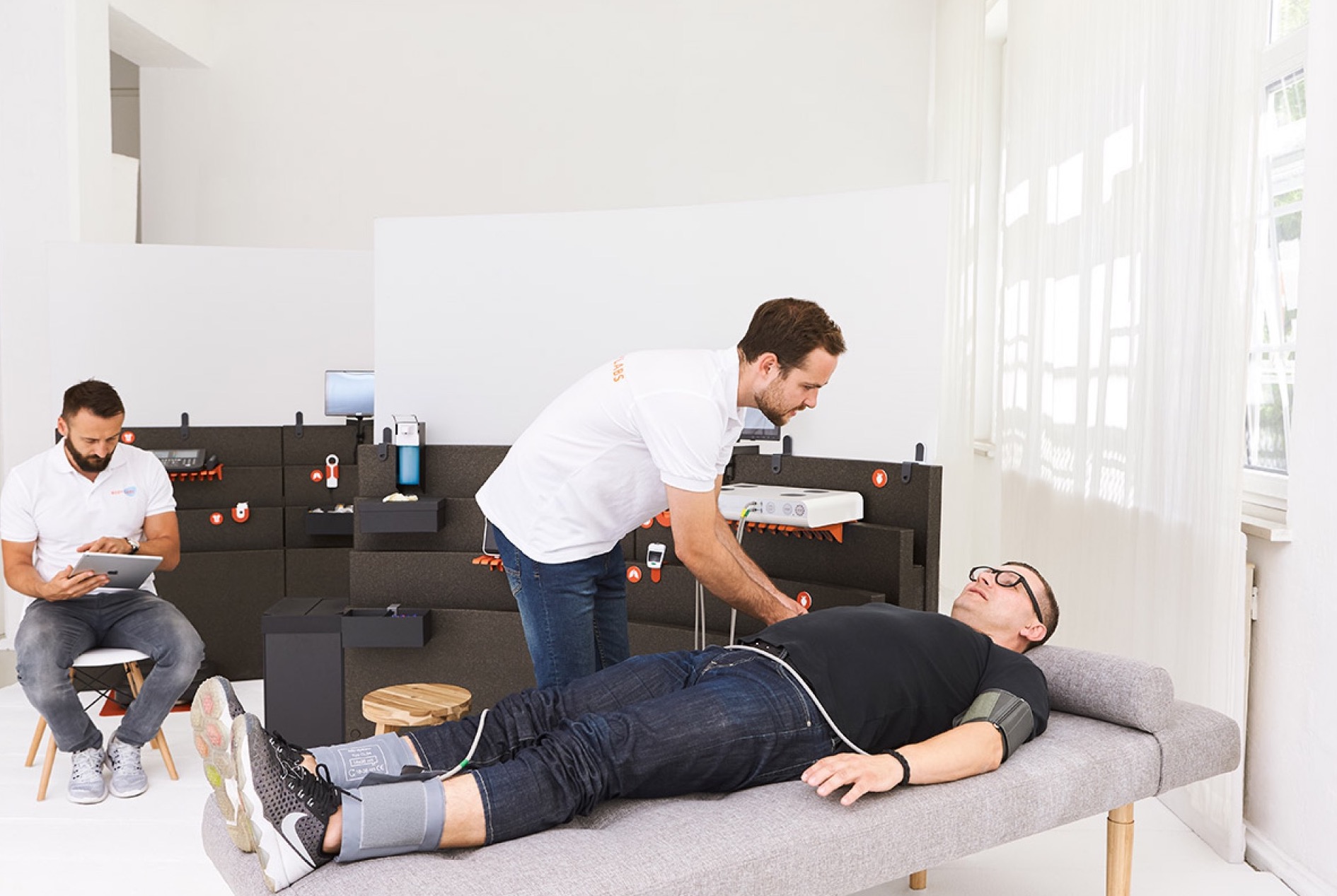 A Bodylabs check-up in a white studio, one person on a bench with a blood-pressure cuff and a technician beside them