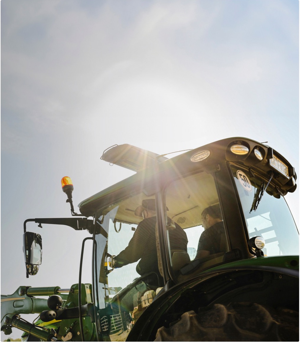 Two people in the cab of a John Deere tractor, seen from below against the sun